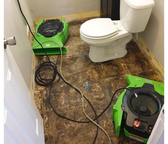 White toilet with two green air movers on the floor. 