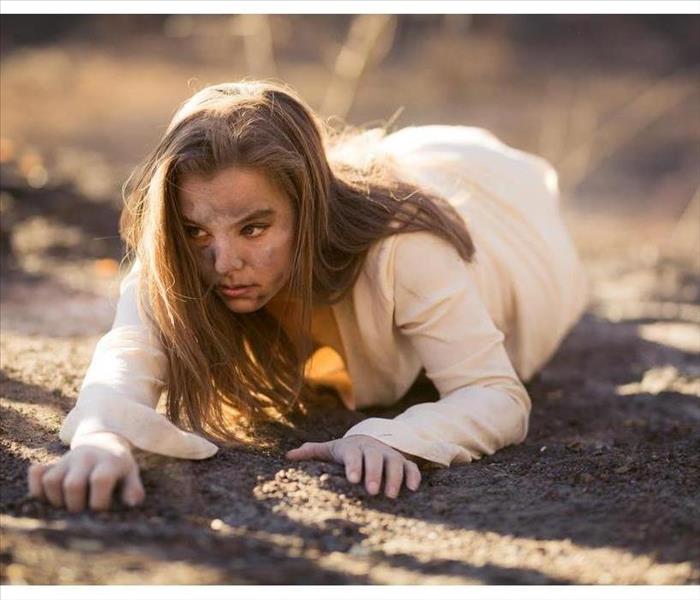 Young mixed race woman in white dress crawls on ground by trees destroyed by wildfire while covered in ashes